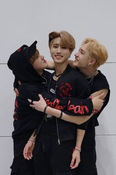 three young men hugging each other in front of a white wall and wearing black clothing