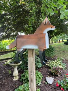 a metal dog statue sitting on top of a wooden post next to flowers and trees