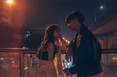 a man standing next to a woman on top of a balcony at night with city lights in the background