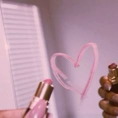 a person holding a pink lipstick with a heart drawn on the wall in the background