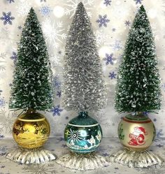 three small christmas trees sitting in vases on a table with snowflakes behind them