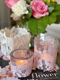 two glass candles sitting on top of a table next to pink flowers and a book