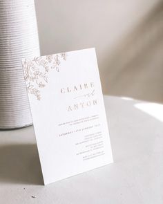 a white and gold wedding card sitting on top of a table next to a vase