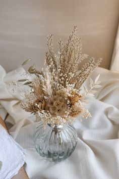 a glass vase filled with dried flowers on top of a white bed sheet covered in sheets