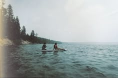 two people are sitting on a surfboard in the water near some trees and sand