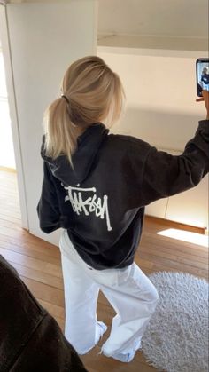 a woman taking a selfie with her cell phone while wearing a black sweatshirt and white pants