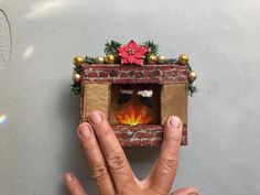 a hand is holding up a miniature fire place in the middle of a brick wall