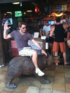 a man riding on the back of a brown bear statue in a bar with people standing around