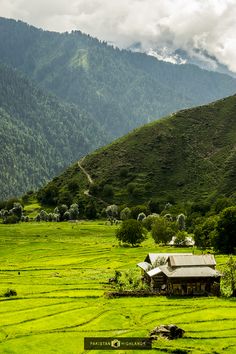 a house in the middle of a lush green field with mountains in the background and clouds overhead