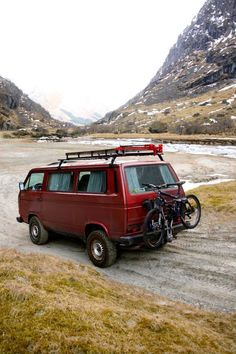 a red van parked on top of a dirt road next to a bike rack and snow covered mountains