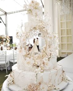 a white wedding cake with flowers on it and a bride and groom in the background