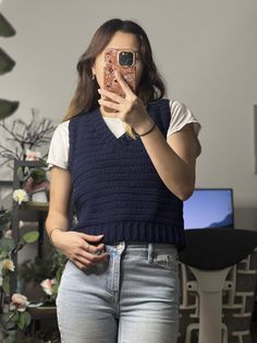 a woman taking a selfie with her cell phone in front of her face while wearing jeans and a sweater vest