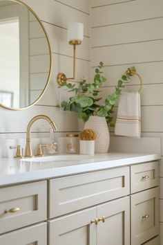 a bathroom with white cabinets and gold accessories on the counter top, along with a round mirror