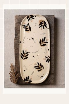 a white plate with black leaves on it sitting on top of a wooden board next to a plant