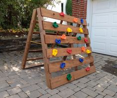 a wooden climbing frame with colorful rocks on it in front of a brick building and garage