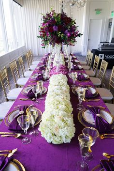 a long table is set with purple and white flowers, gold rimmed plates and silverware