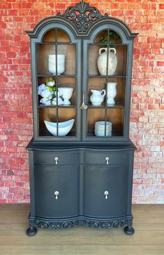 an old china cabinet with glass doors and flowers on top is displayed in front of a brick wall