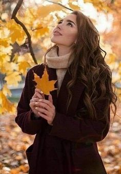 a woman standing in front of a tree holding an orange maple leaf and looking up at the sky