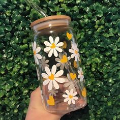 a hand holding a clear glass with white and yellow daisies on the inside, in front of a green hedge