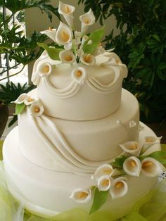 a wedding cake with white flowers and green leaves on the top is sitting on a table in front of a potted plant