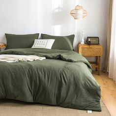 a bed with green sheets and pillows in a white room next to a lamp on a table