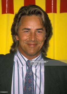 a man with long hair wearing a suit and tie smiling in front of a yellow background