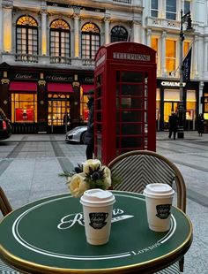 two cups of coffee sit on an outdoor table in front of a phone booth and telephone booth