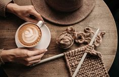 a person holding a cup of coffee on top of a wooden table next to knitted items