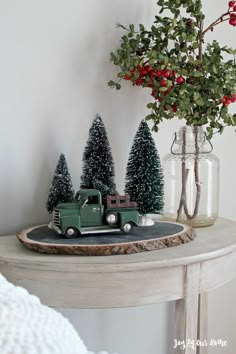 a green truck is parked in front of some christmas trees on a table with a vase full of red berries