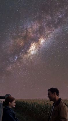 a man and woman looking at the stars in the night sky over a cornfield