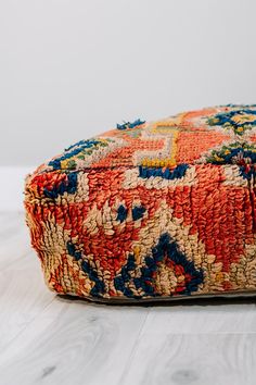 an orange, blue and yellow decorative cushion on a white wooden floor in front of a gray wall