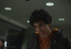 a man with curly hair wearing an orange shirt and black jacket looking at the camera