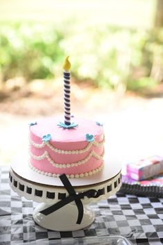 a pink birthday cake with a single candle on it sitting on a checkered table cloth