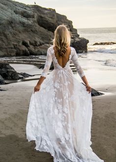 a woman is walking on the beach wearing a white wedding dress with long sleeves and open back