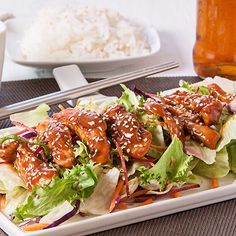 a white plate topped with lettuce covered in sauce next to rice and chopsticks