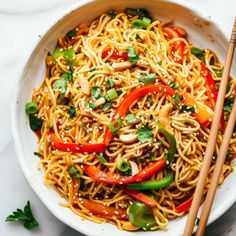 a white bowl filled with noodles and vegetables next to chopsticks on a marble surface