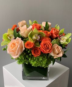 an arrangement of orange and green flowers in a square glass vase on a white table