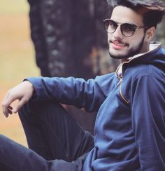 a man sitting on the ground wearing sunglasses and a blue hoodie with trees in the background