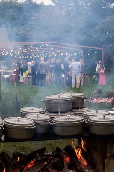 people are gathered around an open fire pit with pots on it and grilling pans in the foreground