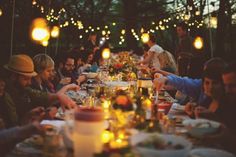 a group of people sitting around a table with food and candles on it at night