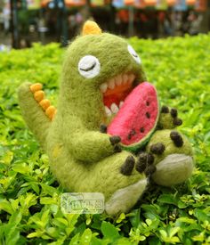 a stuffed toy dinosaur holding a watermelon in its mouth on some green grass