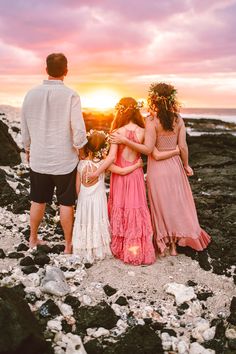 three people standing on rocks near the ocean at sunset with their arms around each other