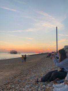 two people sitting on the beach at sunset taking pictures with their cell phones and one person walking by