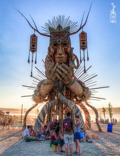 a group of people standing in front of a giant statue on the beach with many sticks sticking out of it's face