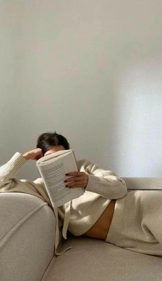 a woman sitting on a couch reading a book with her legs crossed and head propped up