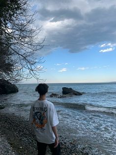 a person standing on the beach looking out at the water and rocks under a cloudy sky