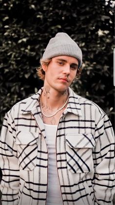 a young man wearing a plaid jacket and beanie standing in front of a tree