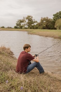 a young man sitting on the bank of a lake while holding onto a fishing rod