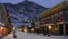 a snowy street with buildings and lights on each side in front of a snow covered mountain