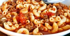 a white bowl filled with pasta and meat on top of a wooden table next to a spoon